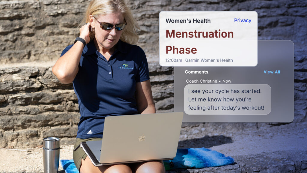 This image features a coach (blonde woman in sunglasses and a dark blue polo shirt) sitting outside, looking at her silver laptop. Overlaying the right side of the image is a screenshot of the Women&rsquo;s Health Metric Card in TrainingPeaks, it&rsquo;s titled: "Women's Health" with "Privacy" in the top right. Below this, it reads "Menstruation Phase" in large red font, with "12:00 am Garmin Women's Health" underneath. Further down, under "Comments," a message from "Coach Christine" (timestamped "Now") reads: "I see your cycle has started. Let me know how you're feeling after today's workout!" The overall scene suggests a coach monitoring an athlete's health data.