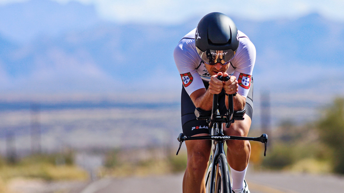 Triathlete Completes An Aerobic Threshold Coupling Workout Fit Male Triathlete On Bicycle With Aerobars Rides Down A Road With Mountains In The Background