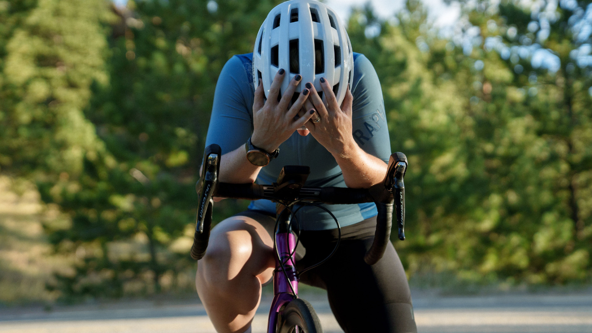 Cyclist Standing Over Her Bike After A Hard Ride, With Her Head Down And Hands On Her White Helmet.